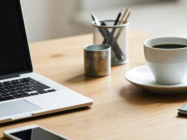 Minimalist desk with laptop, notebook and coffee cup near sunlit window