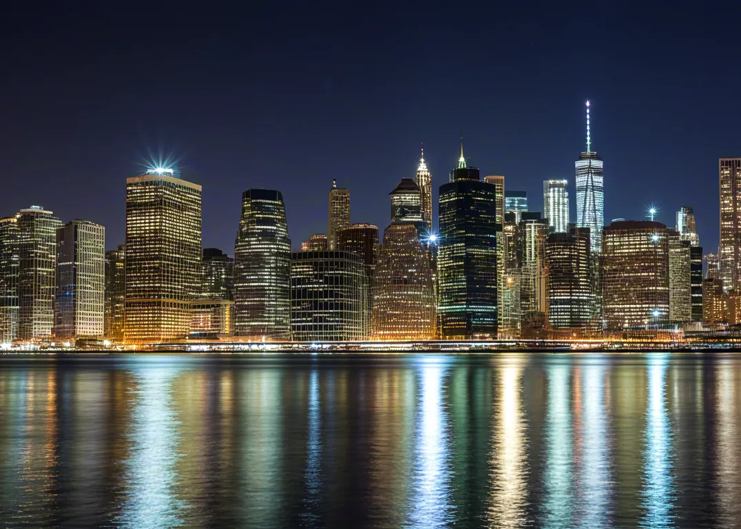 Modern city lights reflecting on calm river under night sky.