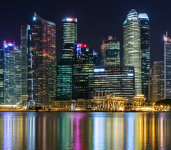 Urban skyline at night with multicolored reflections on calm surface.