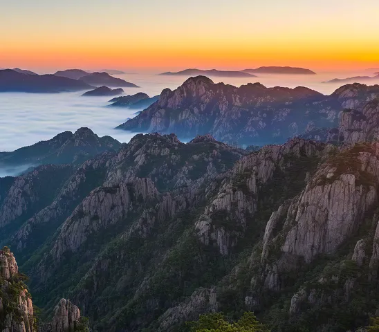 Mountains covered in fog at dawn with warm orange sunlight on horizon.