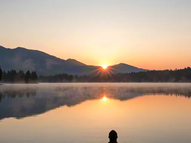 Person meditating in lotus position by a calm lake during sunrise, surrounded by mountains and mist, symbolizing peace, mindfulness, and harmony with nature