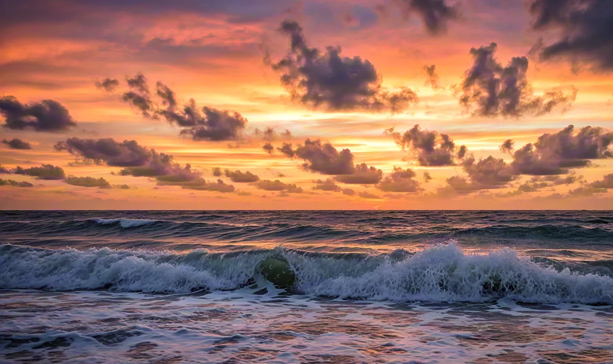 Scenic view of ocean waves rolling under a vibrant sunset sky with orange and purple clouds.